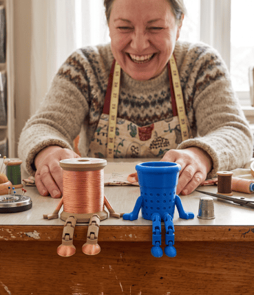 Colorful 3D printed robot figures on a craft table with a smiling woman. Acadiana3DPrints